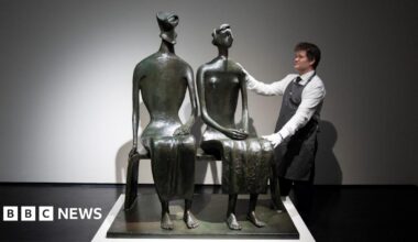 A bronze sculpture of a couple sitting on a bench. A man in white shirt and apron poses with his hands on the sculpture