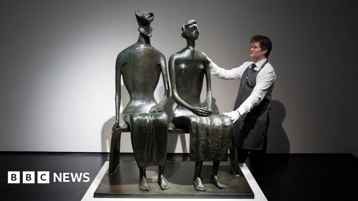 A bronze sculpture of a couple sitting on a bench. A man in white shirt and apron poses with his hands on the sculpture