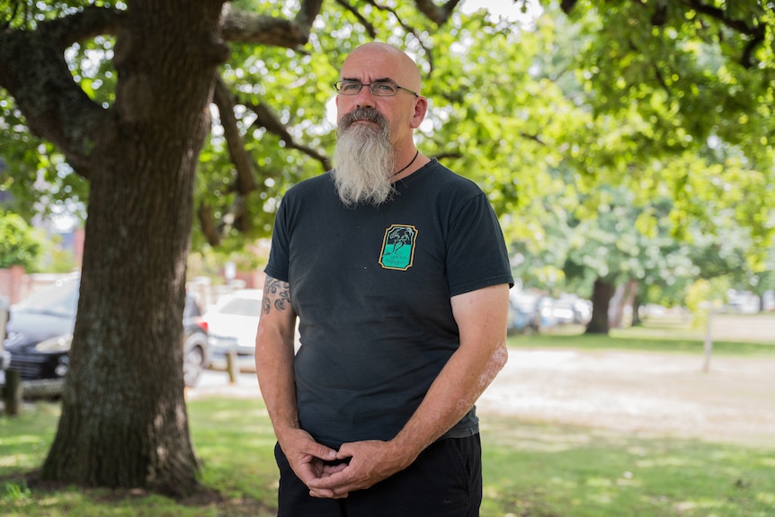 Scott standing in a park. He has a beard and tattoos on his arm.