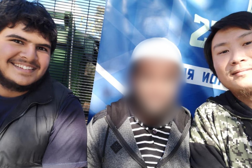 A photo of three people. The young men on either side smile, the middle person's face is blurred.