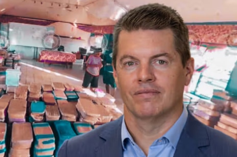 A man with short, dark hair wears a dark suit while standing aboard what appears to be a ship.