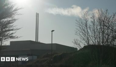 The Beddington Energy Recovery Facility is seen from outside, with two tall chimneys releasing a plume of steam into the sky, surrounded by trees and fencing.