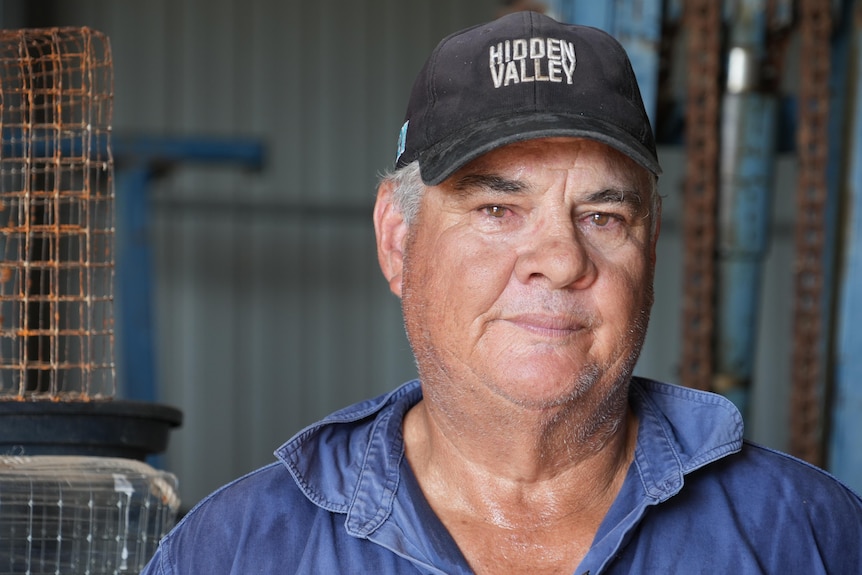 Garry Warrick looks at the camera. He is wearing a black cap and blue polo shirt. He is standing in a shed.