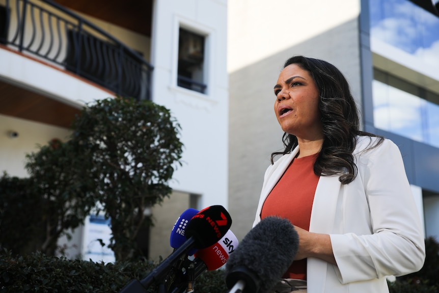 A side-on shot of Jacinta Nampijinpa Price speaking into microphones during a media conference outside, wearing a white jacket.