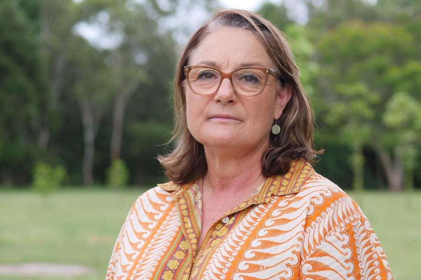 A woman in front of a green park, she is wearing an orange patterned shirt and glasses