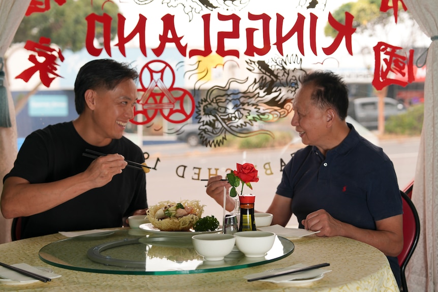 Two men siting on one side of a round table holding chopsticks. In front of them is a cooked dish on a plate.