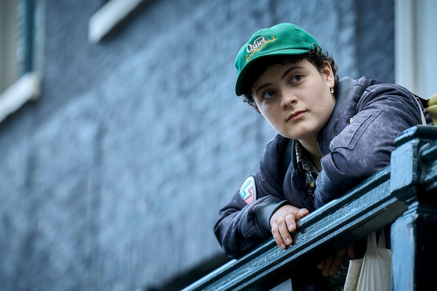 Person with short curly hairs wears a green cap and leans over a railing with brick facade behind them