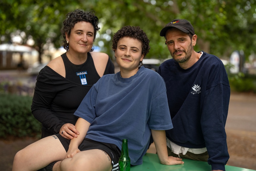 A young person with short, curly hair seated and slightly smiling. Standing is woman with black cut-out top and man in a cap