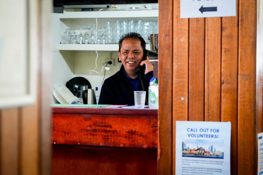 A dark haired man in a black top holds a phone to his ear as he stands behind a counter with glasses stacked on a shelf behind.