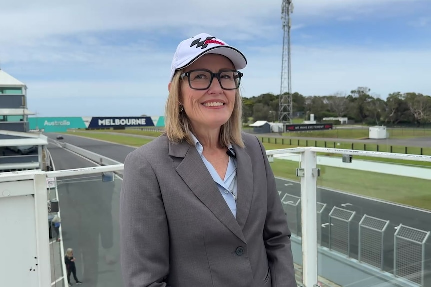 A woman with blonde hair and glasses is wearing a motoGP cap, standing alongside the race track.