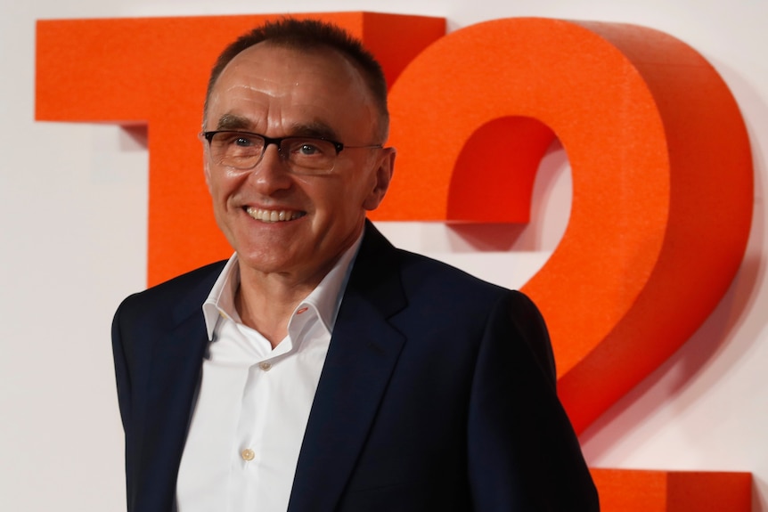 Older male film director in glasses and blue suit smiles and poses on red carpet. Big orange T2 sign in back.