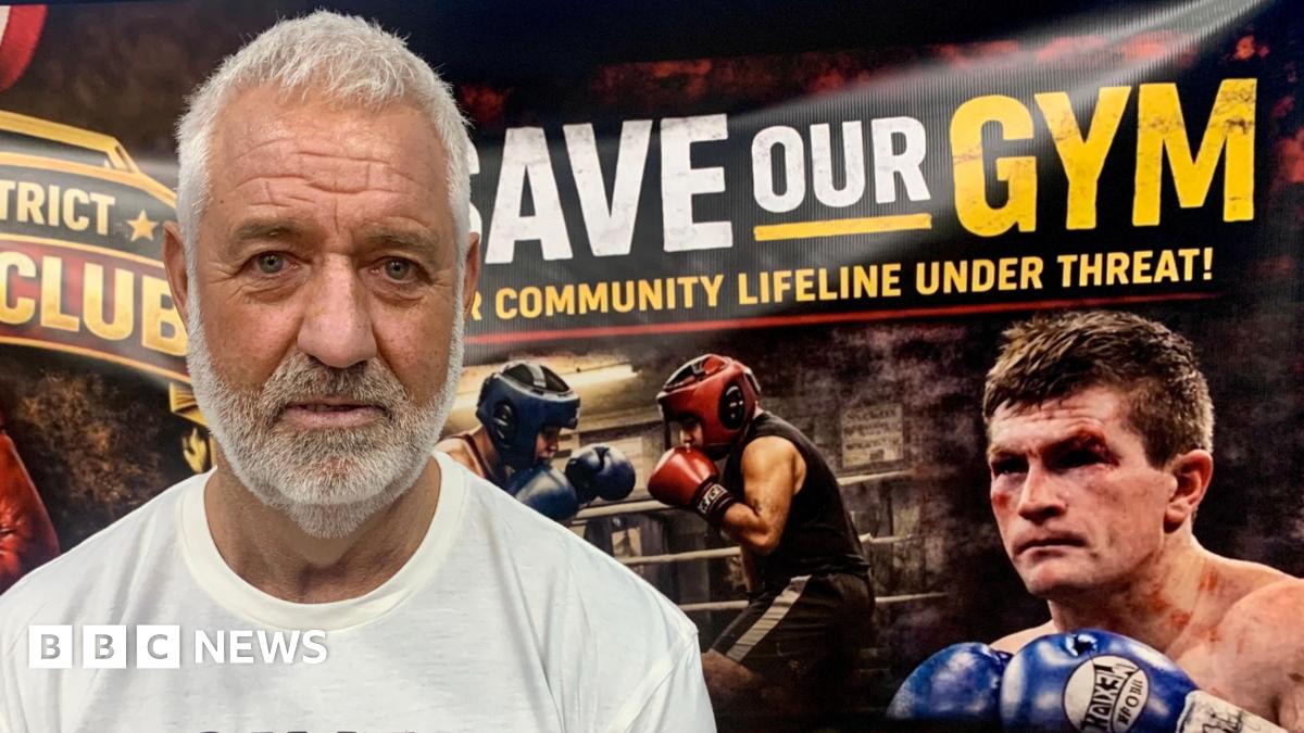 Steve Strong has white hair and a matching beard. He is wearing a white t-shirt and stands in front of a "Save Our Gym" banner which includes a photograph of two amateur boxers sparring, and a bloodied Ricky Hatton from one of his fights.