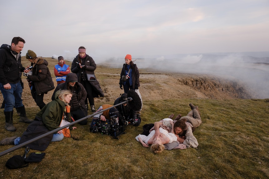 Two people lay on a grassy mountain surrounded by a filming crew.