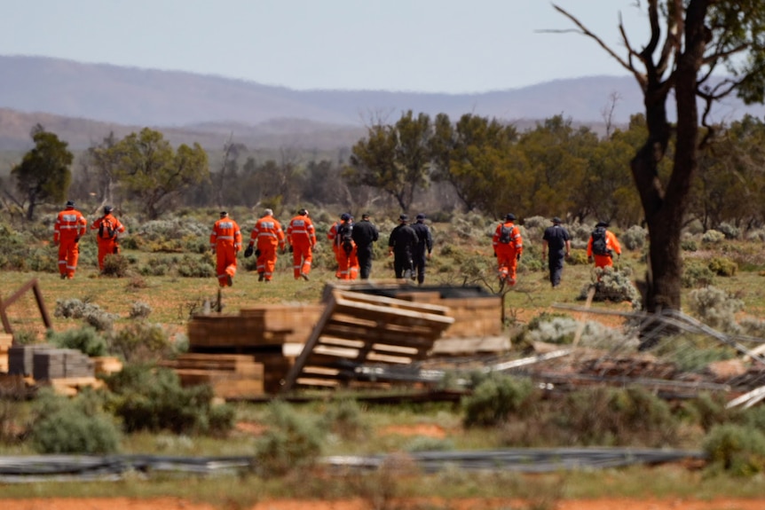 A row of SES volunteers in orange and police officers in navy uniforms walk across arid ground 