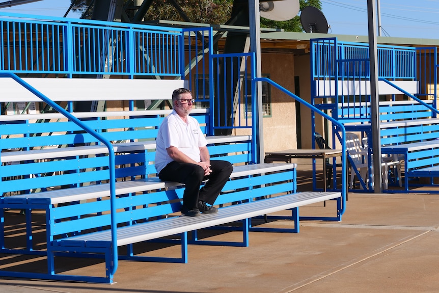 Middle aged man in white shirt and black pants sits in empty blue stands.