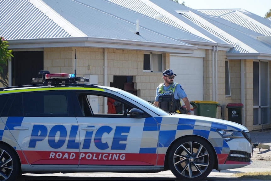 A policeman stands next to a police car parked in front of a house with a hole in the wall