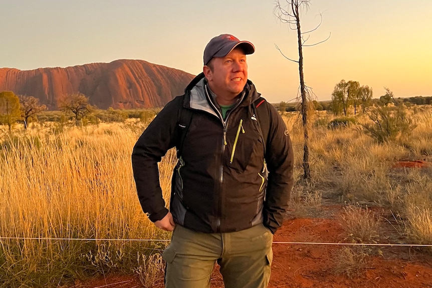 A man wearing a jacket and cap, standing in front of Uluru.