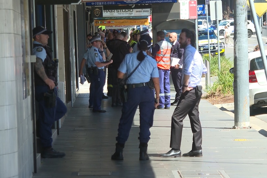 Police officers stand on footpath of shopping strip, talking to each other and members of the public
