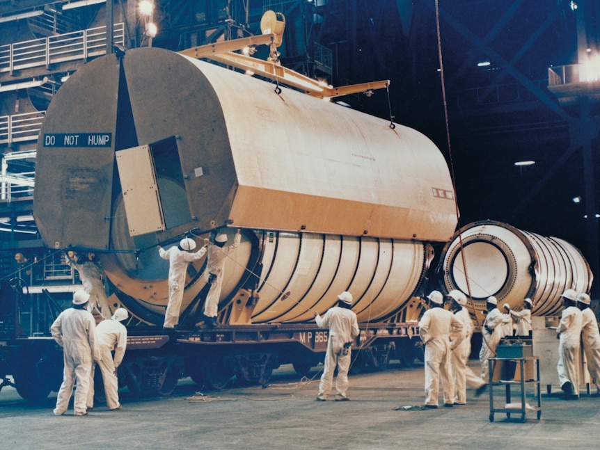 A large cylindrical container is lifted off a white rocket section on a train carriage, surrounded by engineers in overalls.