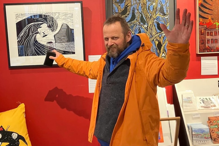 Jon Kudelka stands next to artworks on a wall in a gallery.