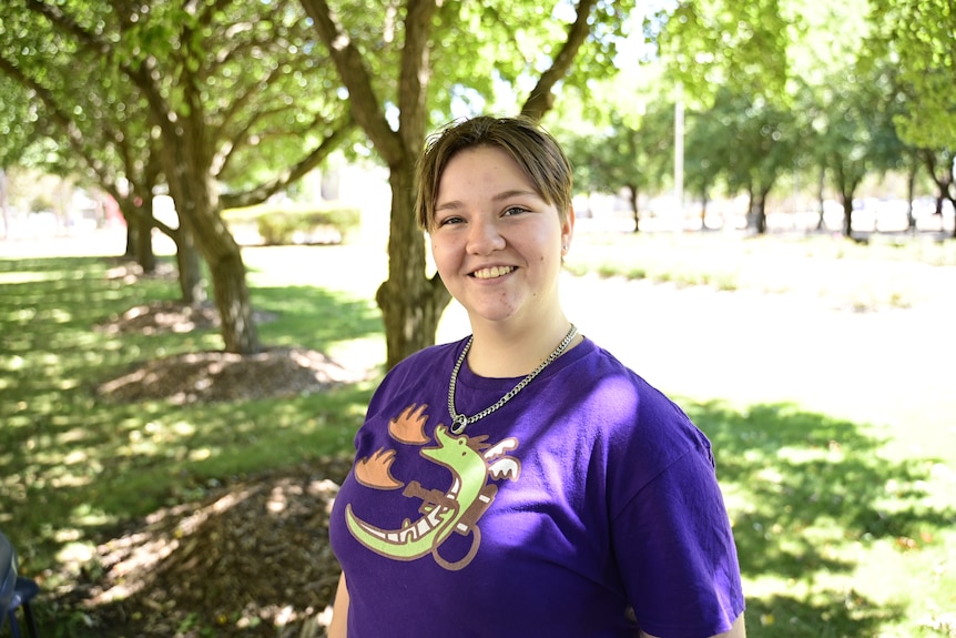 A young person in bright purple t-shirt smiles, trees out of focus in background.