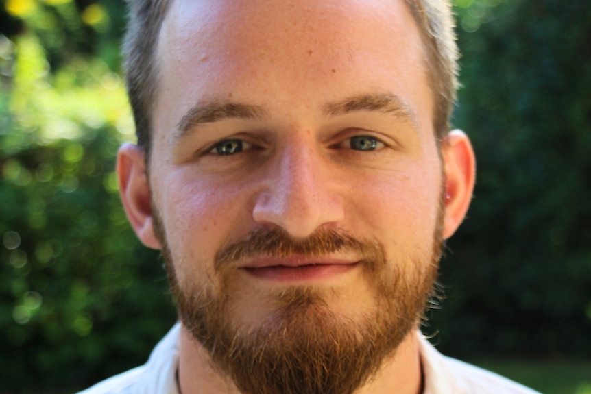 A close up of a man looking at the camera. A green natural background out of focus behind him.
