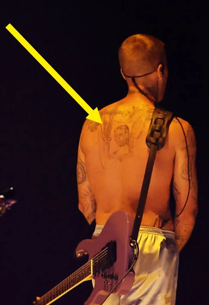 Musician on stage in shorts, holding an electric guitar behind their back. Upper body is tattooed and shirtless