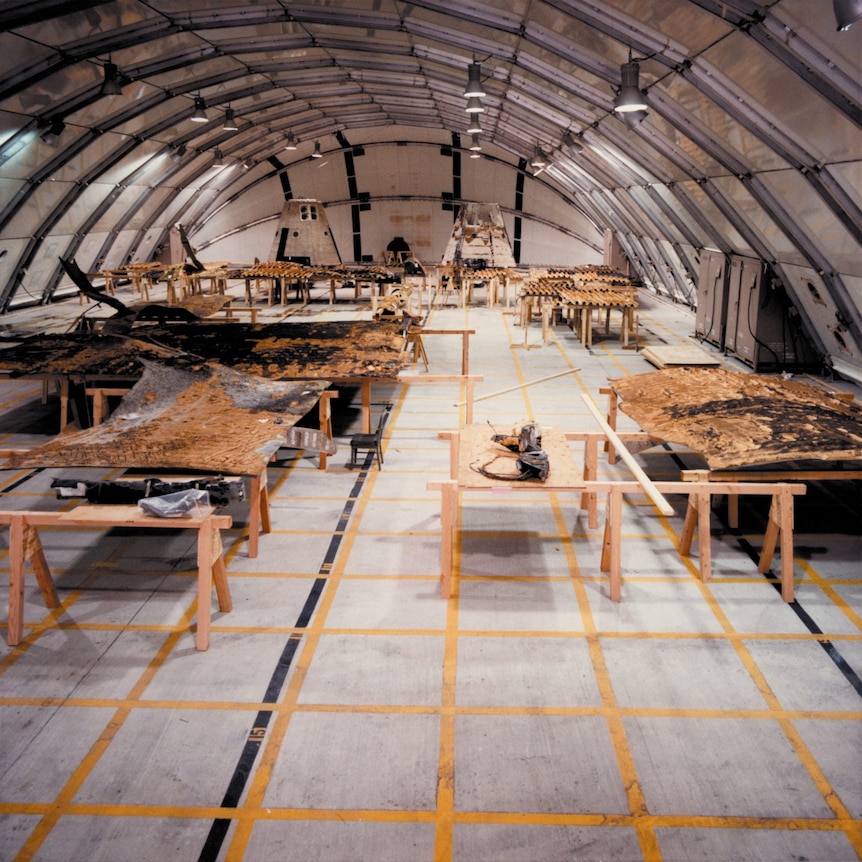 Photo of a large room with a curved roof with a grid marked on the floor in yellow and many objects on trestle tables.