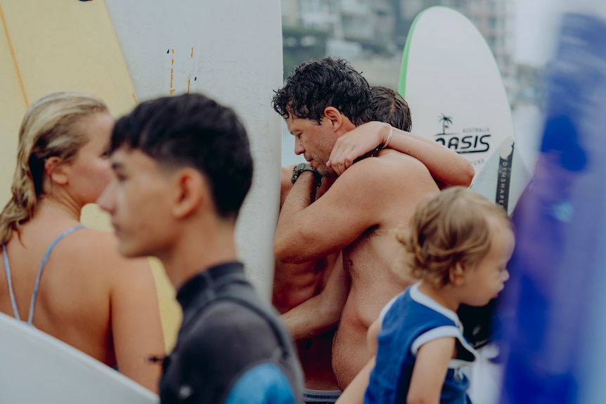 Two men seens embracing each other among a crowd of people with surf boards and paddleboards at the beach.