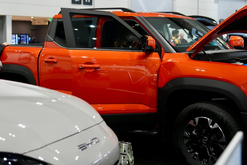 An orange ute with bonnet up and doors open, in a showroom.