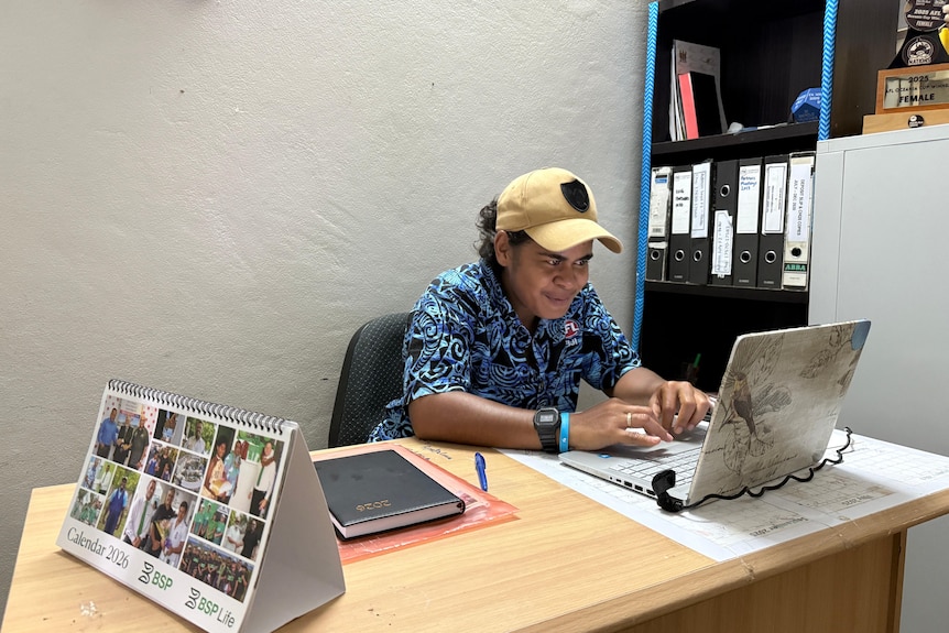 A young lady in a blue button up shirt sits at an office desk working on a laptop she is also wearing a fawn cap on her head.