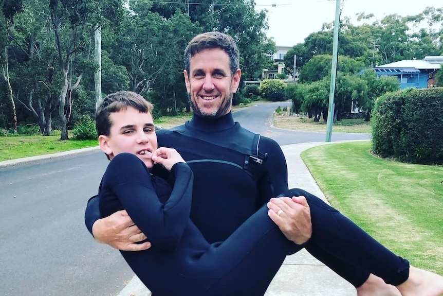A man is carrying his son, both in wet suit