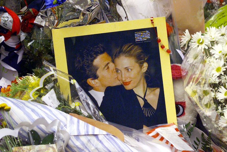 A framed photograph of a dark-haired man and a glamorous blonde woman sits on a pile of floral tributes.