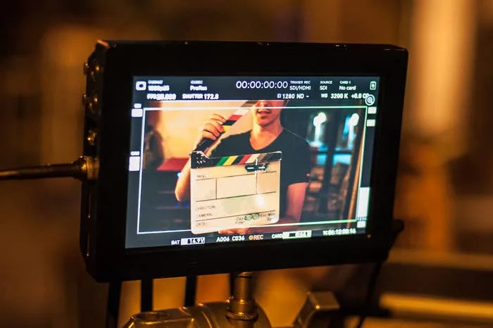 A film monitor displays a person holding a clapperboard on a set, signifying the start of a scene in a TV or movie production