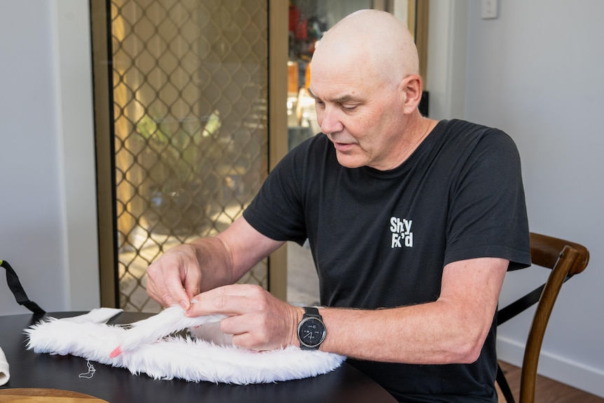 A bald man sitting at a pool table stitching a fake tail together.