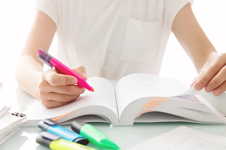 Person highlighting text in a book with a pink marker, surrounded by other colored markers on a desk