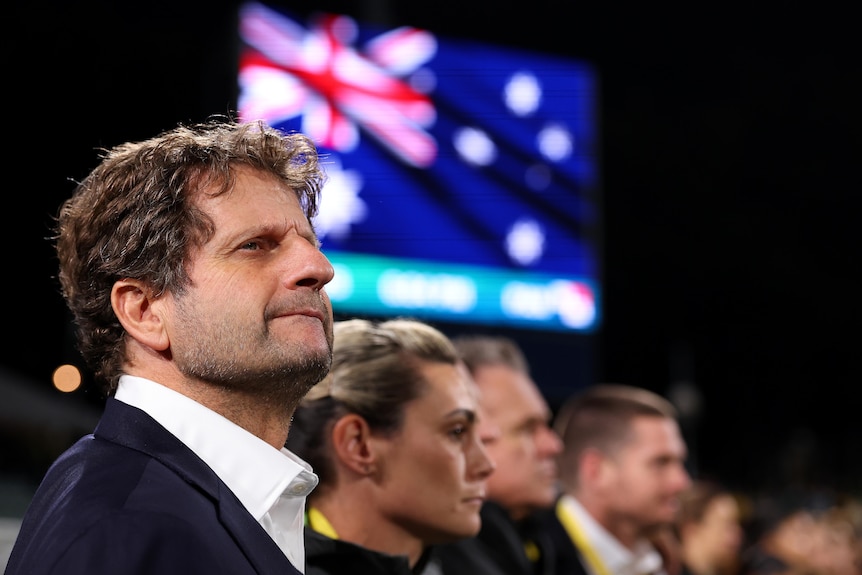 A close up of Matildas coach Joe Montemurro's face with the Australian flag on a big screen in the background