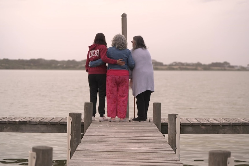 Three women stand at the end of a jetty which arms around each other. 