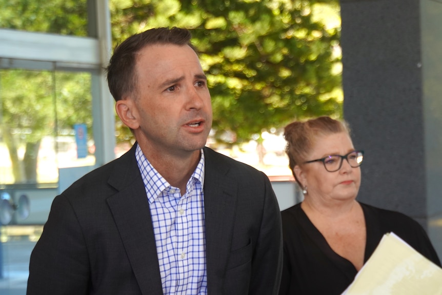 Michael stands at the forefront of the image with a woman standing behind him in the background, he looks off centre and talks.