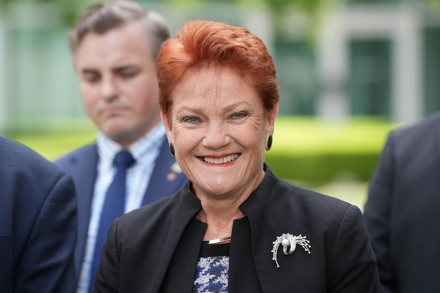 A smiling, red-haired woman — Pauline Hanson — wears a dark blazer with a brooch as she stands outdoors.