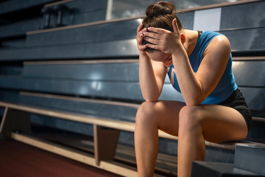 A female gymnast sits on a bench with her head in her hands