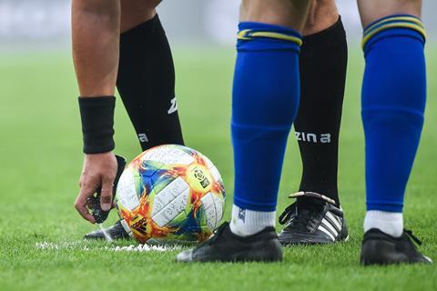 A photo from the knees down of a football referee applies some white spray foam to the pitch in front of a football with a player standing watching.