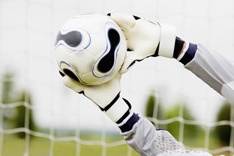 A football goalkeeper wearing protective gloves stretching to block a football from going into the goal