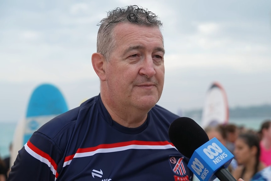 Mr Jackson wearing a rugby shirt with surf boards and people around him on a beach.