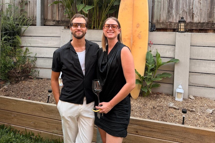 A man and woman stand smiling in a backyard, both wearing black clothes and sunglasses