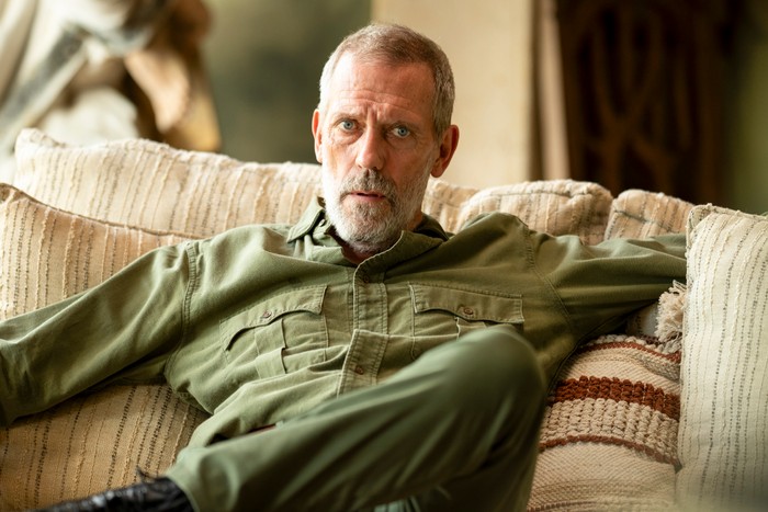 Richard Roper sits on a sofa in his home, wearing a green shirt and trousers, staring ahead with concern