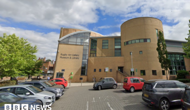 A modern, light-coloured brick building with lettering on the side which indicates it houses an art gallery, museum and library. There is a car park in the foreground of the image with car parks in rows on either side. There are a number of trees nearby the building.