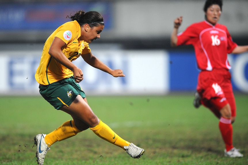 A soccer in green and gold kicks a ball with a player in red behind her