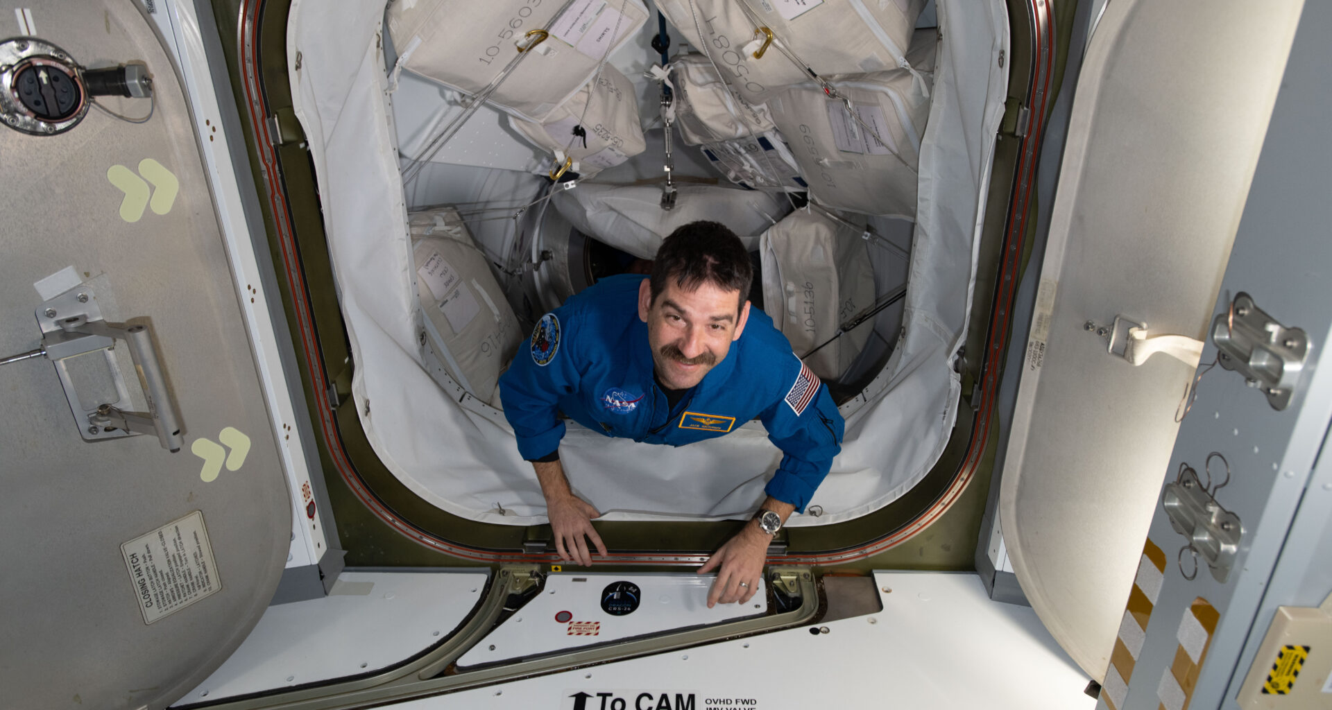 A man with dark hair and a mustache looks up at the camera, smiling. His hands are on the bottom edge of the hatch entrance as he makes his way inside the space station. The path he is coming through has many square cargo bags strapped to each side.