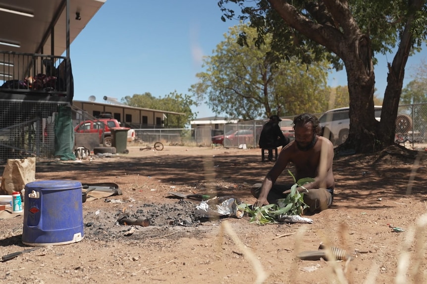 A man sitting in front of an unlit campfire, in a large yard of bare earth, in a remote community.